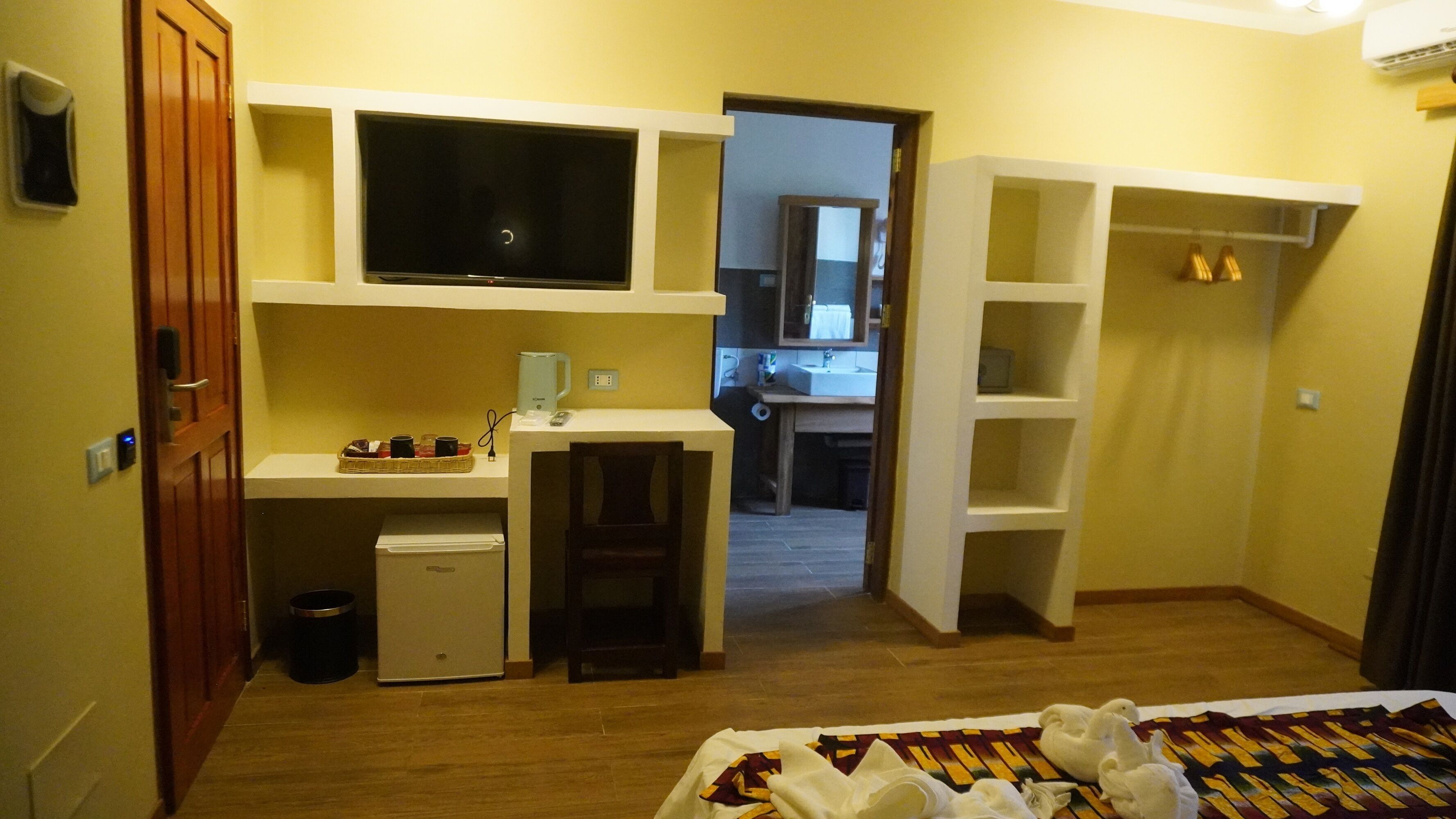 Habitación de hotel pequeña con kitchenette, televisión, refrigerador compacto, escritorio y cama con colcha estampada.