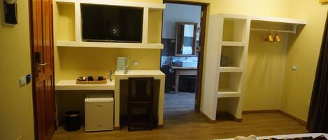 Habitación de hotel pequeña con kitchenette, televisión, refrigerador compacto, escritorio y cama con colcha estampada.