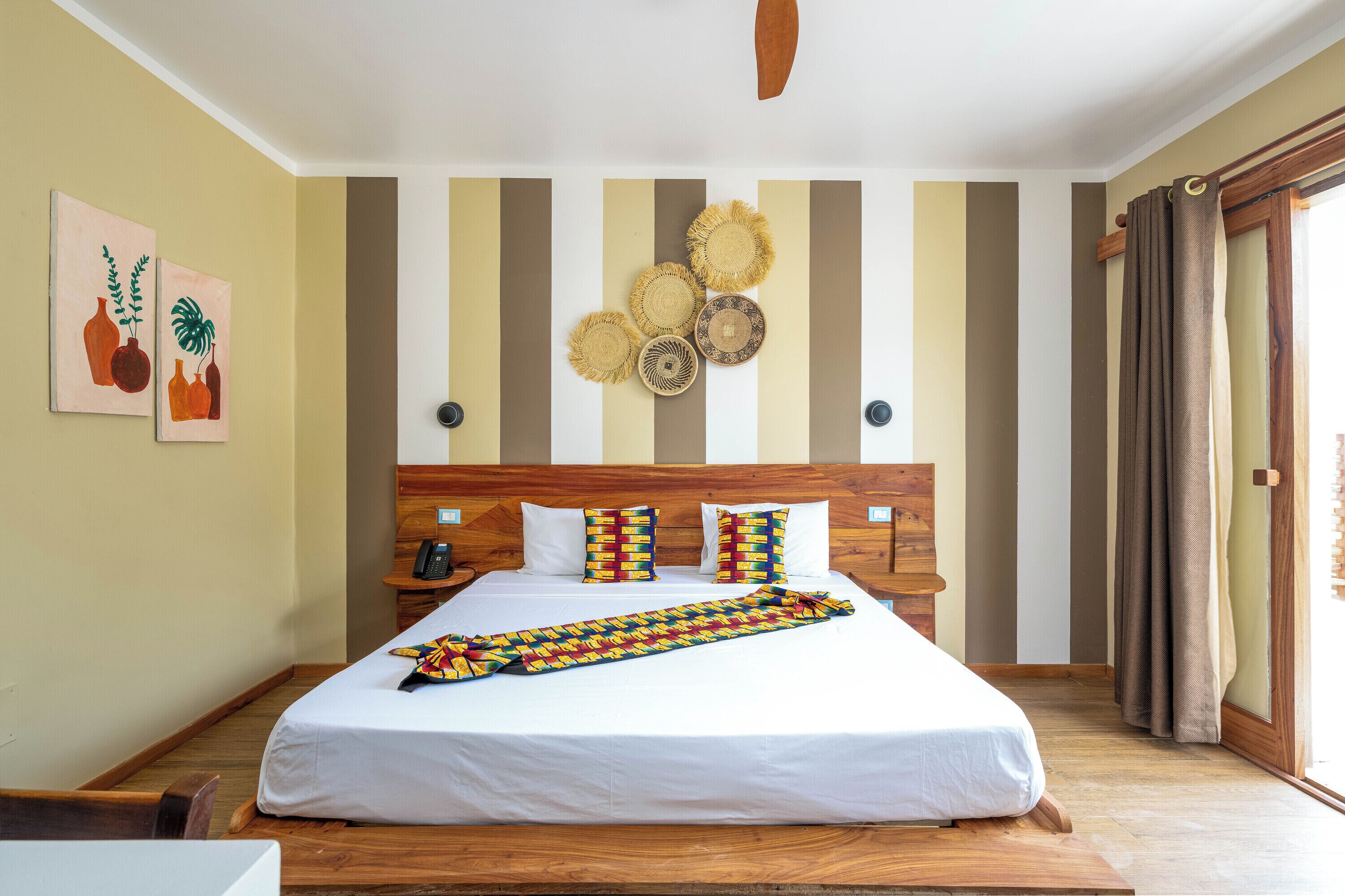 Une chambre d’hôtel avec un lit, une tête de lit en bois, un décor mural à rayures et une fenêtre avec des rideaux.