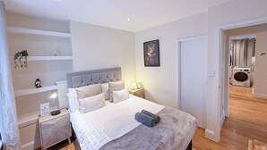 A bedroom with a bed, bedside tables, a nightstand, a wall-mounted shelf, a framed picture, and a door leading to a bathroom with a washing machine.