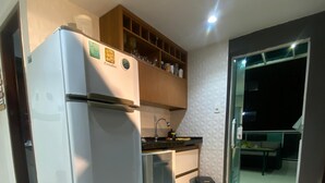 Een compacte keuken met een koelkast, een gootsteen en een klein aanrecht. Een prullenbak en een deur naar een balkon.