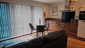 A modern living room with a sofa, a glass dining table with chairs, a kitchen area with a stove and oven, and a large window with blinds. - 2nd Floor City Apartment (Sheffield)
