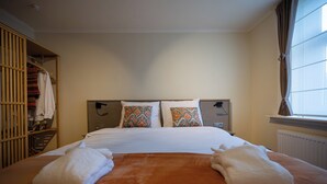 Ein ordentlich bezogenes Bett mit zwei Handtüchern, ein Kleiderschrank mit Kleidung und ein Fenster mit Vorhängen.