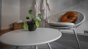 Ein runder, weißer Tisch mit einer grünen Pflanze im schwarzen Topf, ein Korbstuhl mit orangefarbenem Kissen und ein weißer Bademantel, der an einer Wand hängt.