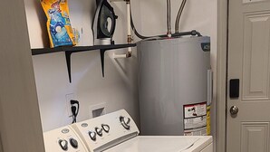 Un cuarto de lavandería con lavadora y secadora de carga frontal, un estante con productos de limpieza y una puerta con picaporte.
