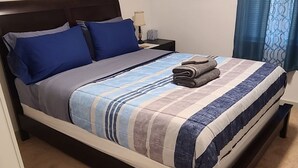 Una cama bien hecha con ropa de cama azul y blanca, una lámpara de noche y un cuadro en la pared.