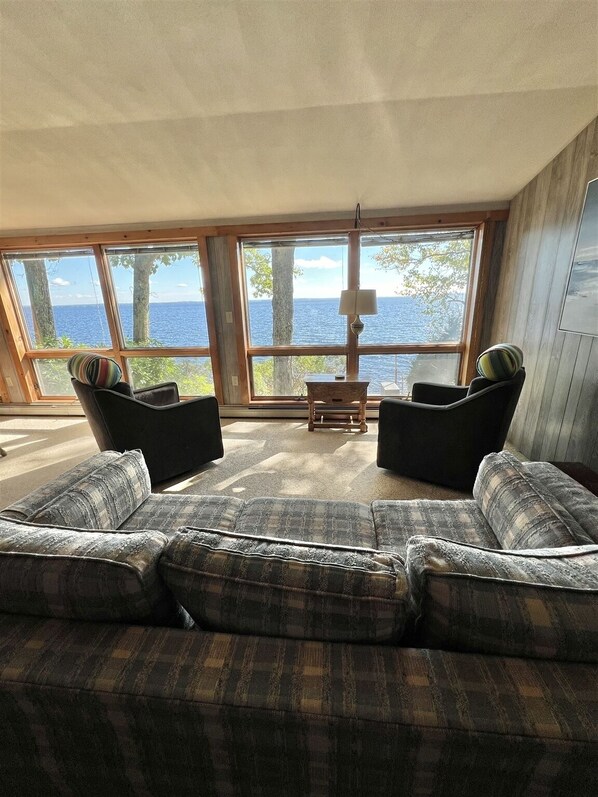 A living room with a large window overlooking a body of water, two black armchairs, a plaid sofa, and a small wooden table. - WATERFRONT 5 BEDROOMS AND 2 BATHROOMS (Sebago)
