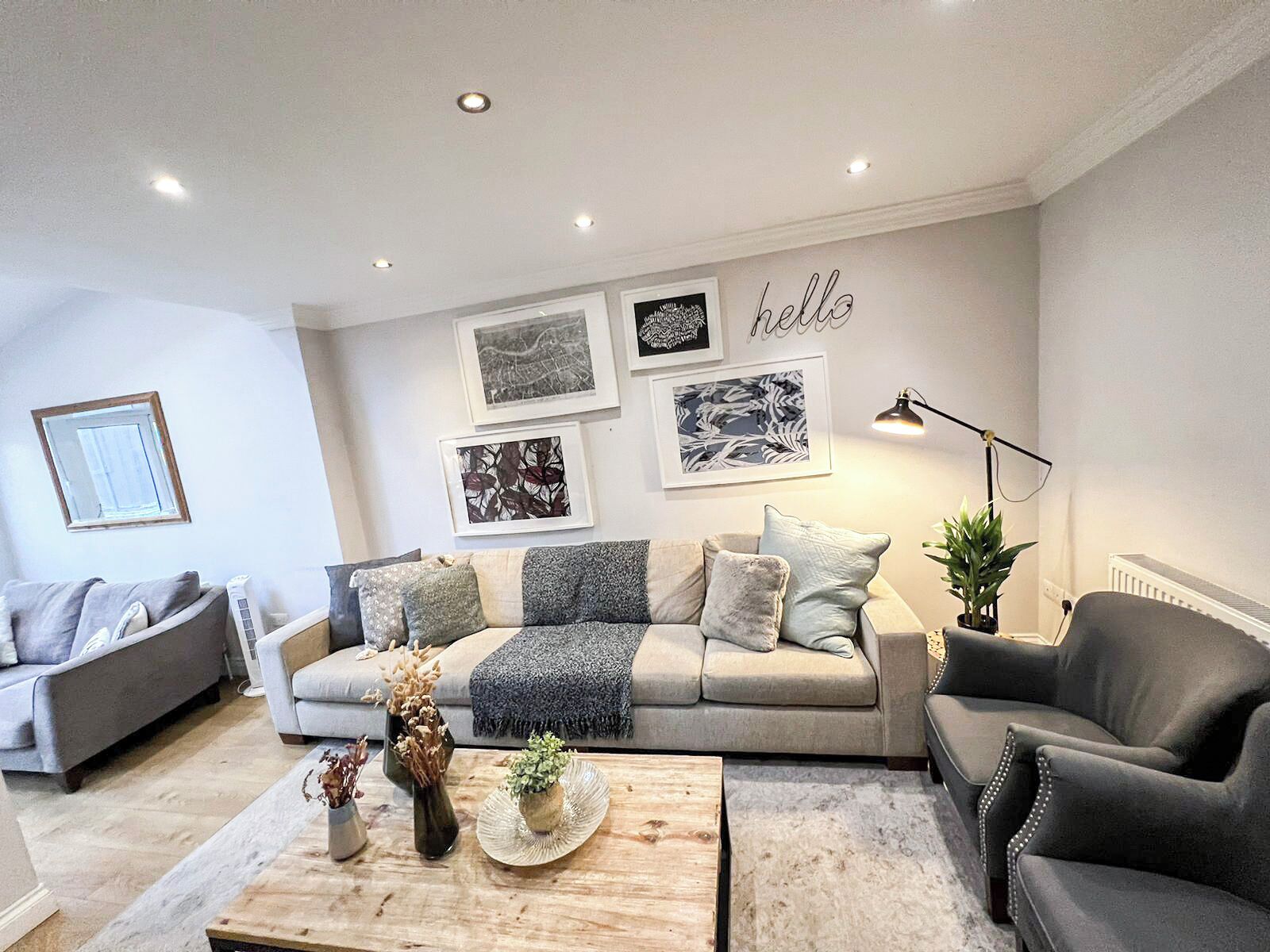 A modern living room with a sofa, two armchairs, a coffee table, and a floor lamp. There are framed pictures on the wall and a 'hello' sign.