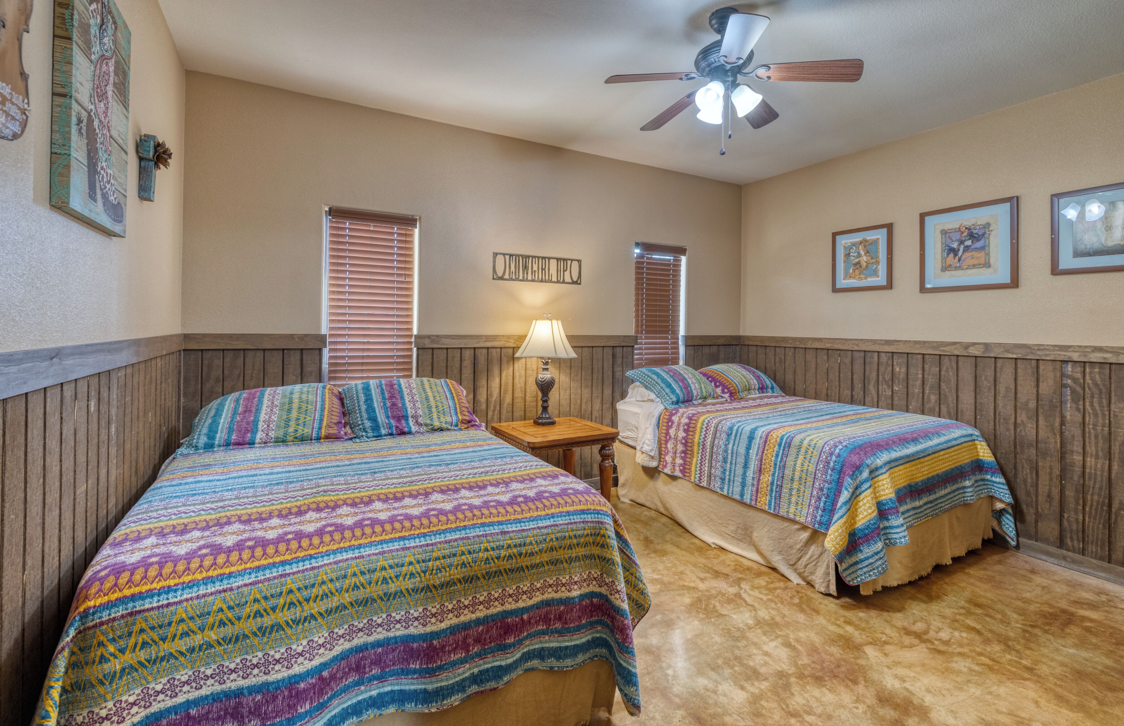 A bedroom with two beds, wooden paneling, a ceiling fan, and framed artwork on the walls.