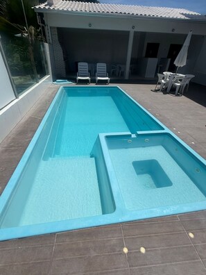 A rectangular swimming pool with a hot tub section, surrounded by a patio area with chairs and tables. - New duplex near Coroa Vermelha beach in Porto Seguro- BA (Santa Cruz Cabrália)