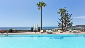 A swimming pool with lounge chairs and palm trees, overlooking a coastal area.