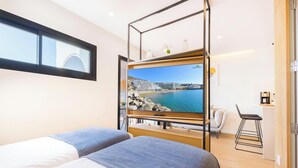 A modern hotel room with a large TV on a wooden stand, two beds, a small seating area, and a view of a coastal town.
