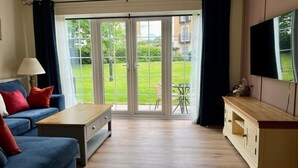 Una sala de estar con un sofá azul, una mesa de centro de madera, un televisor en la pared y un ventanal corredizo que da a un jardín.