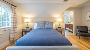A bedroom with a large bed, two bedside tables with lamps, a chair, a window with curtains, and a view of the bathroom.