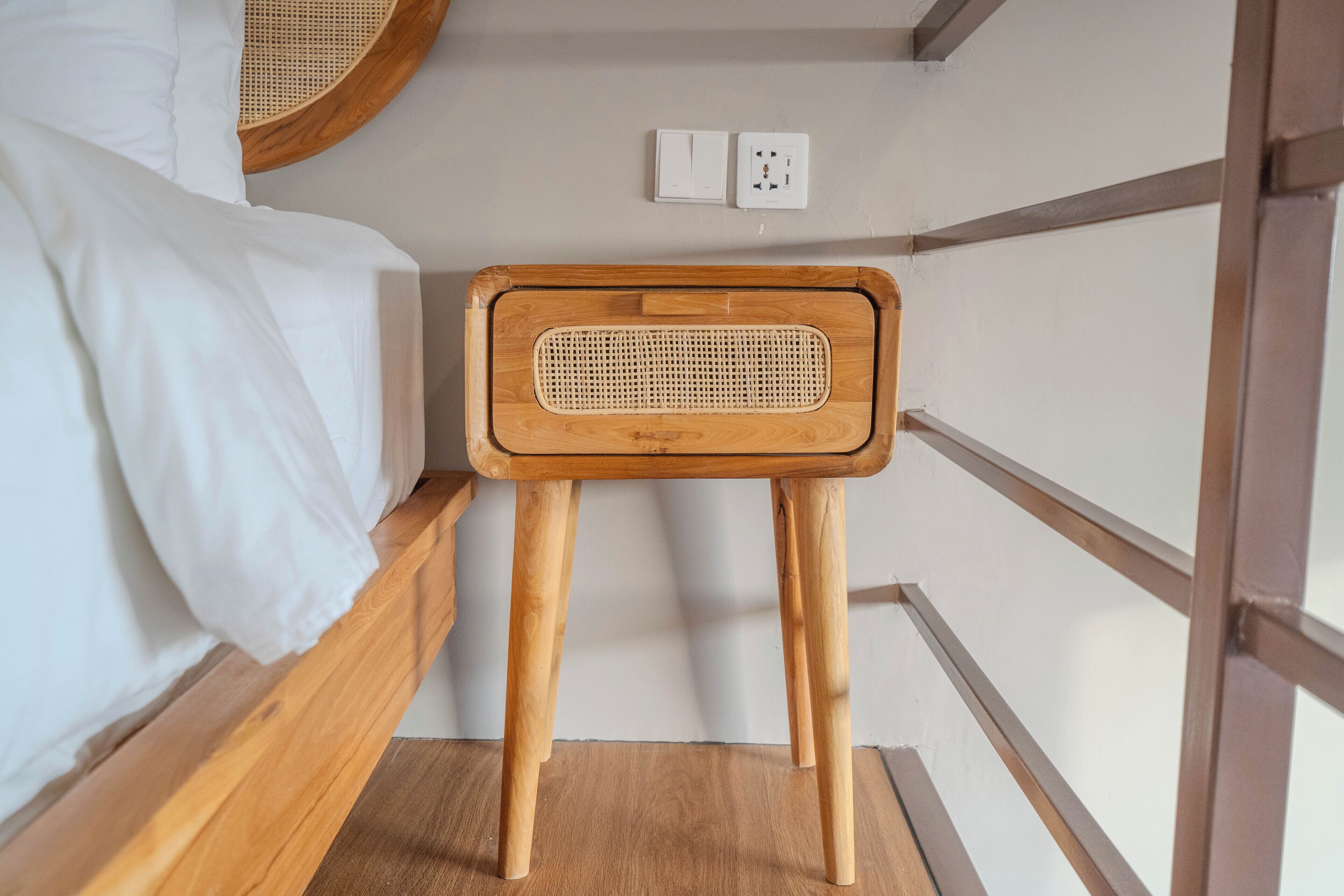 Una stanza piccola con una cuccetta in legno, un tavolino in legno con seduta intrecciata e una presa di corrente.