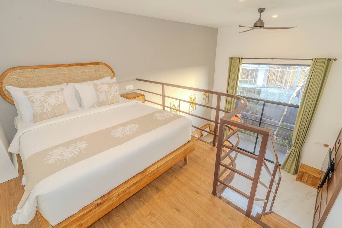 Une chambre avec un lit en bois, un balcon doté d’une rambarde en verre et un ventilateur de plafond.