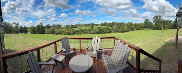 A deck with Adirondack chairs and a table, overlooking a grassy field and trees under a partly cloudy sky.