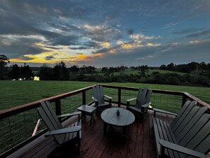 A wooden deck with Adirondack chairs and a table, overlooking a grassy field and a sunset.