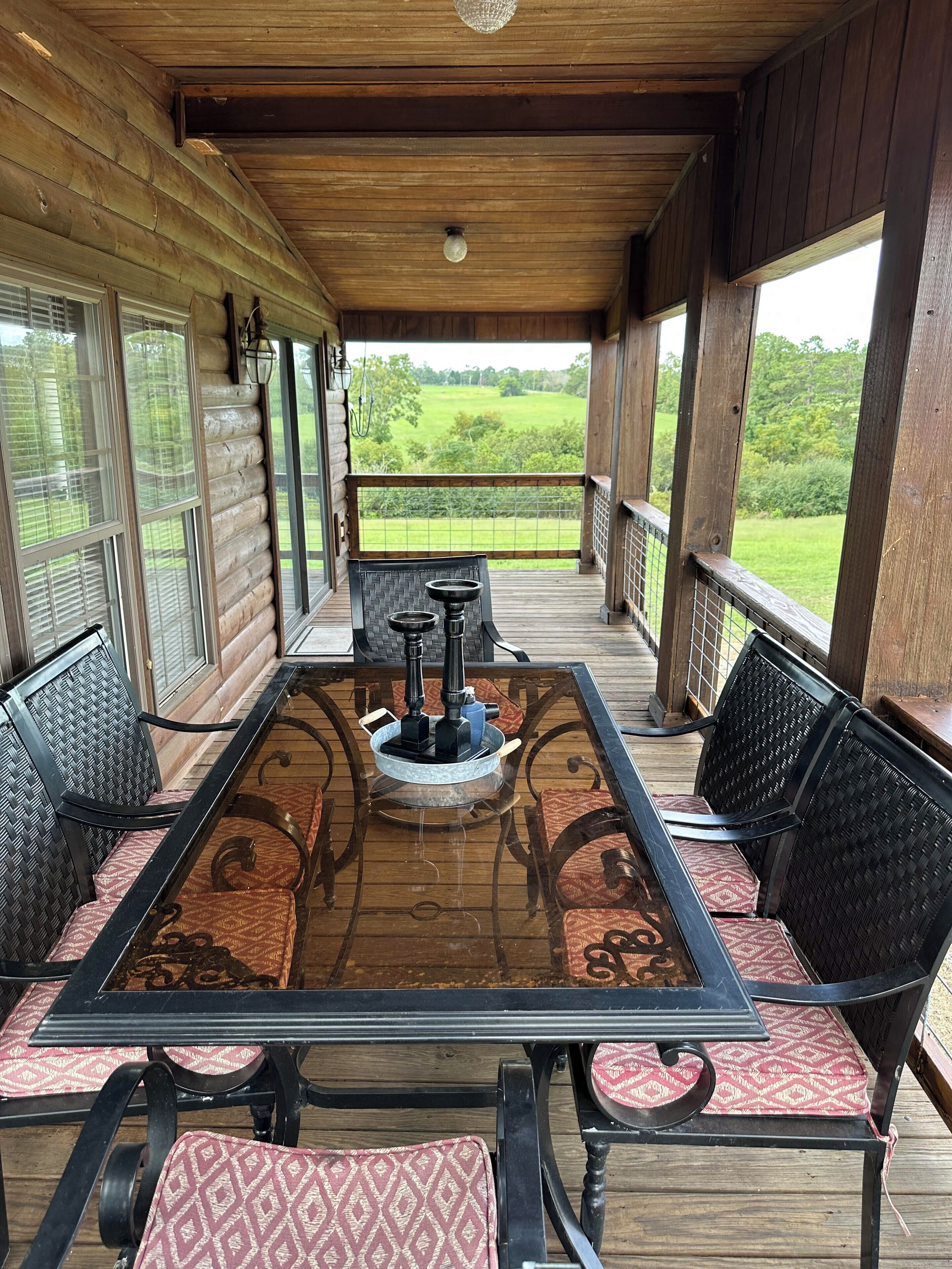 Une terrasse extérieure couverte, dotée d’une table à plateau de verre, de chaises en osier et offrant une vue sur un champ verdoyant.