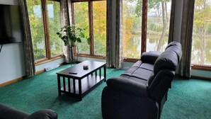 A room with a green carpet, a black sofa, a small table, and a TV on the wall. Large windows offer a view of trees and a body of water. - Dock, Deck and Game Room (Perrysburg)
