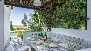 An outdoor dining area with a table set for two, surrounded by white chairs, under a pergola with climbing plants.