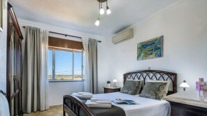 A bedroom with a bed, bedside tables, a window, curtains, an air conditioning unit, and a painting on the wall.