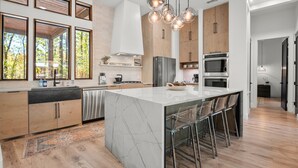 A modern kitchen with a large island, stainless steel appliances, and a view of trees through large windows.