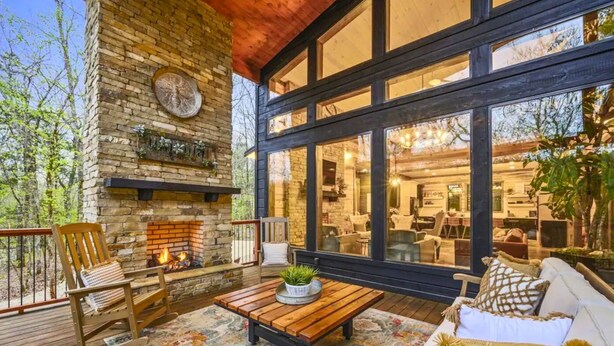 A cozy outdoor patio with a stone fireplace, wooden furniture, and large windows offering views of the interior.