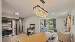 Une cuisine moderne dotée d’appareils électroménagers en acier inoxydable, un coin repas avec une table et des chaises en bois, et un salon avec un canapé et une grande fenêtre.