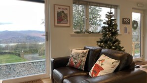 A living room with a leather sofa, a Christmas tree, and a scenic view through the windows. - Swallows Lodge - Modern accommodation with breathtaking views (Keswick)