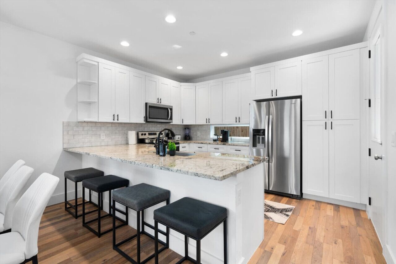 A modern kitchen with white cabinets, a stainless steel refrigerator, a microwave, and a granite countertop. There are three bar stools and a small island with a sink.