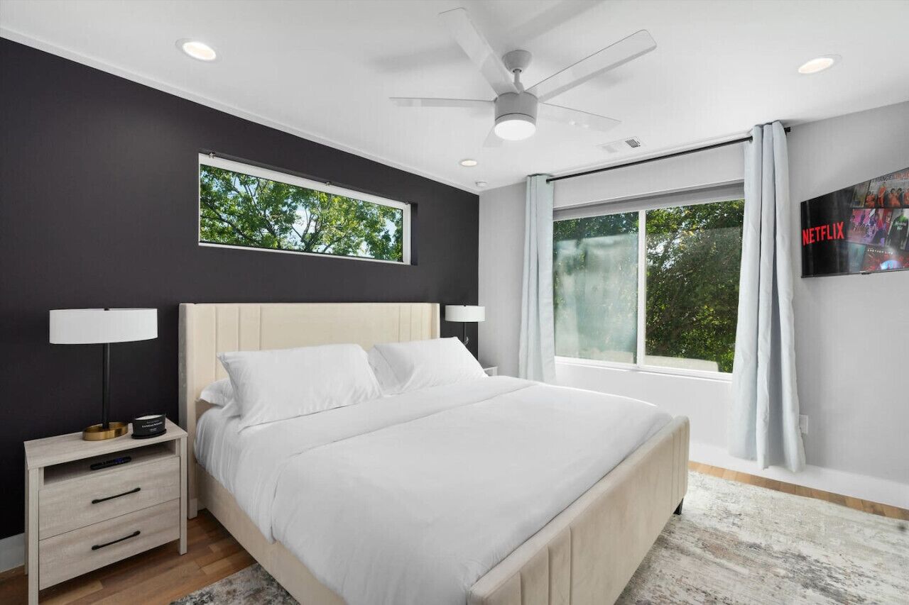 Une chambre à coucher moderne, équipée d’un lit, de tables de chevet, d’un ventilateur de plafond et d’une grande fenêtre avec des rideaux.