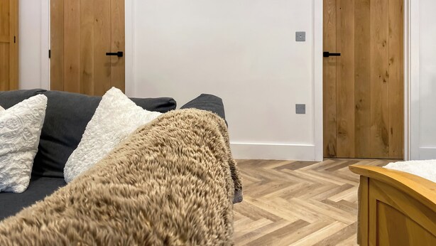 Une pièce avec un plancher en bois, un canapé gris agrémenté de coussins blancs et bruns, et deux portes en bois.