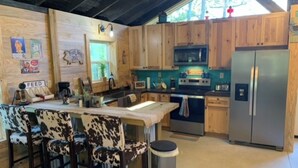 A kitchen with wooden cabinets, a stainless steel refrigerator, a microwave, and a stove. There are bar stools with a cowhide pattern and a trash can. - Charming farmhouse in delightful Brantley with AC (Brantley)