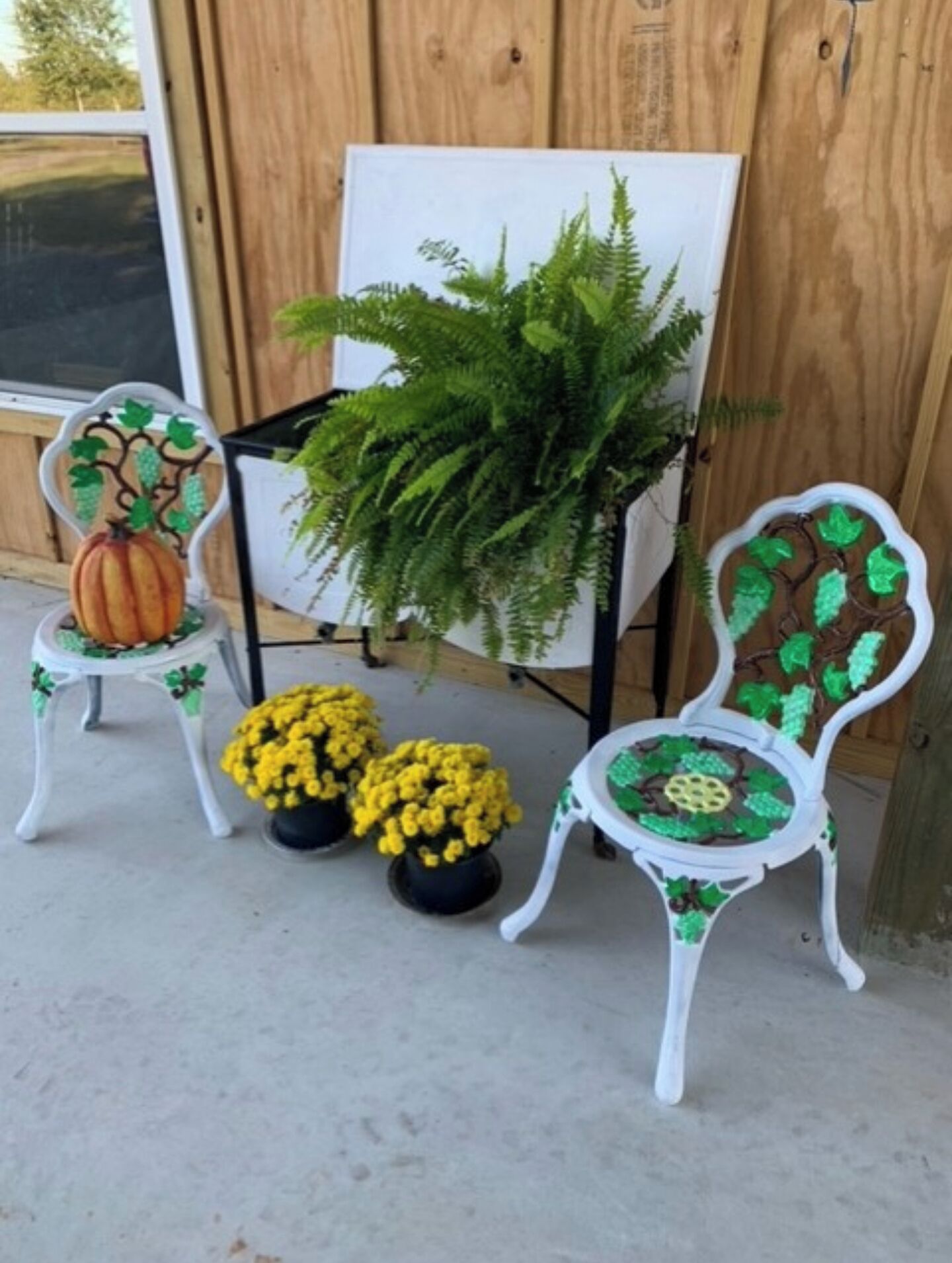 Two white chairs with green leaf designs, a whiteboard, a large green fern, and two potted yellow flowers.