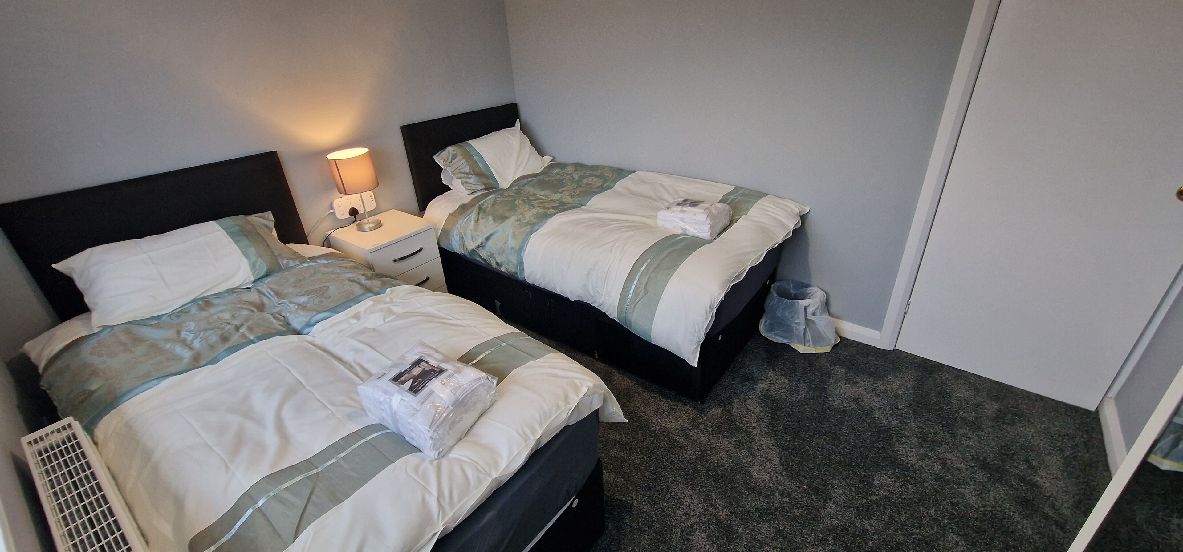A hotel room with two beds, a bedside table with a lamp, a trash bin, and a wall-mounted heater.