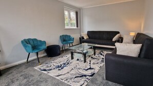 Een woonkamer met een zwarte bank, twee blauwe fauteuils, een glazen salontafel en een kamerbreed tapijt met een patroon.