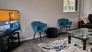 Een woonkamer met een flatscreen-televisie waarop een streamingdienst draait, twee smaragdgroene fauteuils, een zwart pouf en een glazen salontafel.