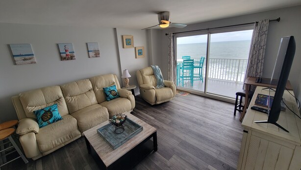 A living room with a beige sofa, two armchairs, a coffee table, and a TV stand. There are framed pictures on the wall, a ceiling fan, and a view of the ocean through large windows.