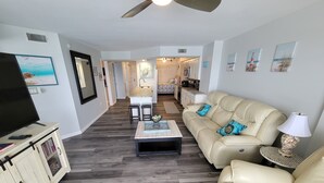 A living room with a beige sofa, a coffee table, and a TV on a cabinet. There is a kitchen in the background.