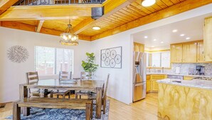 A spacious dining area with a wooden table and chairs, a chandelier, and a view of the kitchen with wooden cabinets and a stainless steel refrigerator.
