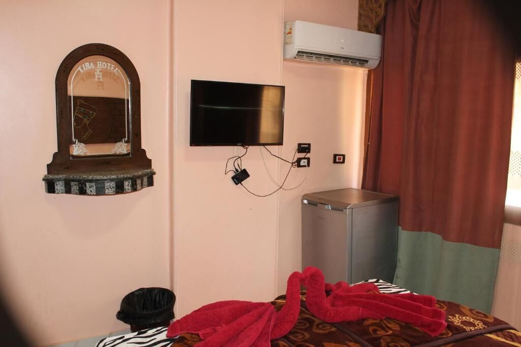 Habitación de hotel con una televisión montada en la pared, un equipo de aire acondicionado pequeño encima y una cama con una colcha estampada y una toalla roja.