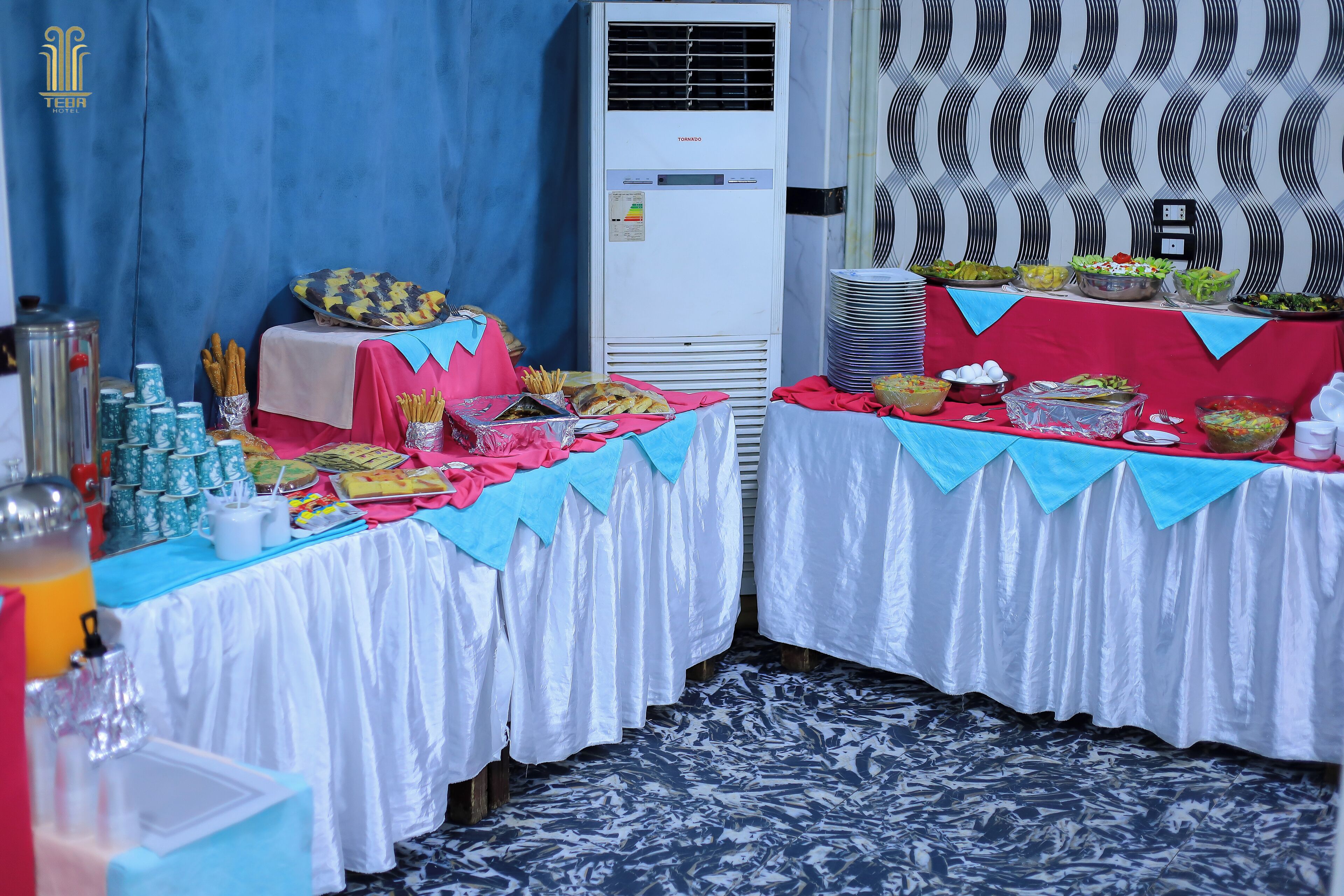A buffet setup with various food items and beverages, a blue curtain, and an air conditioning unit in the background.