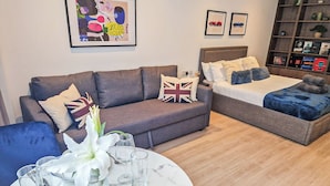 Ein modernes Wohnzimmer mit Sofa, Bett, Esstisch gedeckt mit Tellern, Gläsern und Besteck. An der Wand hängen gerahmte Bilder und ein Bücherregal mit Büchern und Dekorationsobjekten.