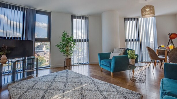 Una sala de estar moderna con un sofá, una mesa de comedor y un televisor. Amplias ventanas con persianas, una planta en maceta y vistas a la ciudad.