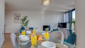 Una sala de estar moderna con una mesa de vidrio dispuesta para el desayuno, un televisor al fondo y un ventanal con cortinas.