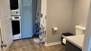 A compact bathroom with a wooden floor, a toilet, a shower curtain, and a washing machine. - Cozy Private Basement in Mid-Town Lincoln (Lincoln)