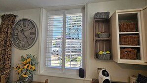 Una habitación con un reloj de pared grande, una ventana con persianas, cestas de mimbre en estantes y un jarrón con girasoles.