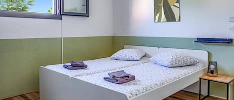 A single-bed room with a wooden floor, a window, a bedside table with a lamp, and a painting on the wall.