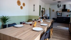 A dining area with a long wooden table set for a meal, flanked by chairs, with a kitchen in the background. - Villa Du Soleil by Villa Plus (Laroque-des-Albères)